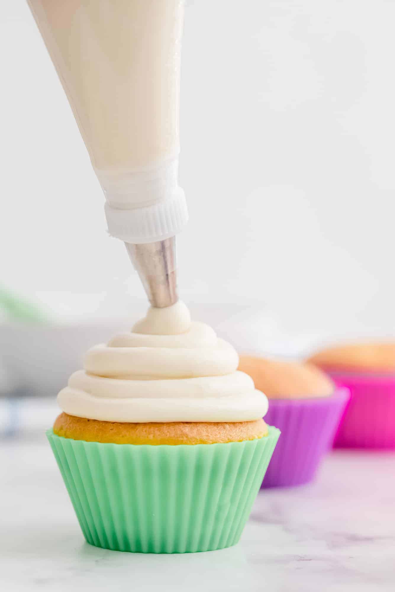 Cream cheese frosting being piped onto a cupcake with a colorful green liner.