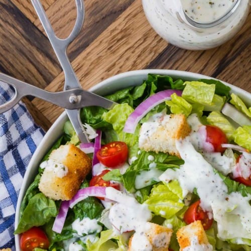 Salad in a bowl with tongs, small jar of italian dressing next to it.