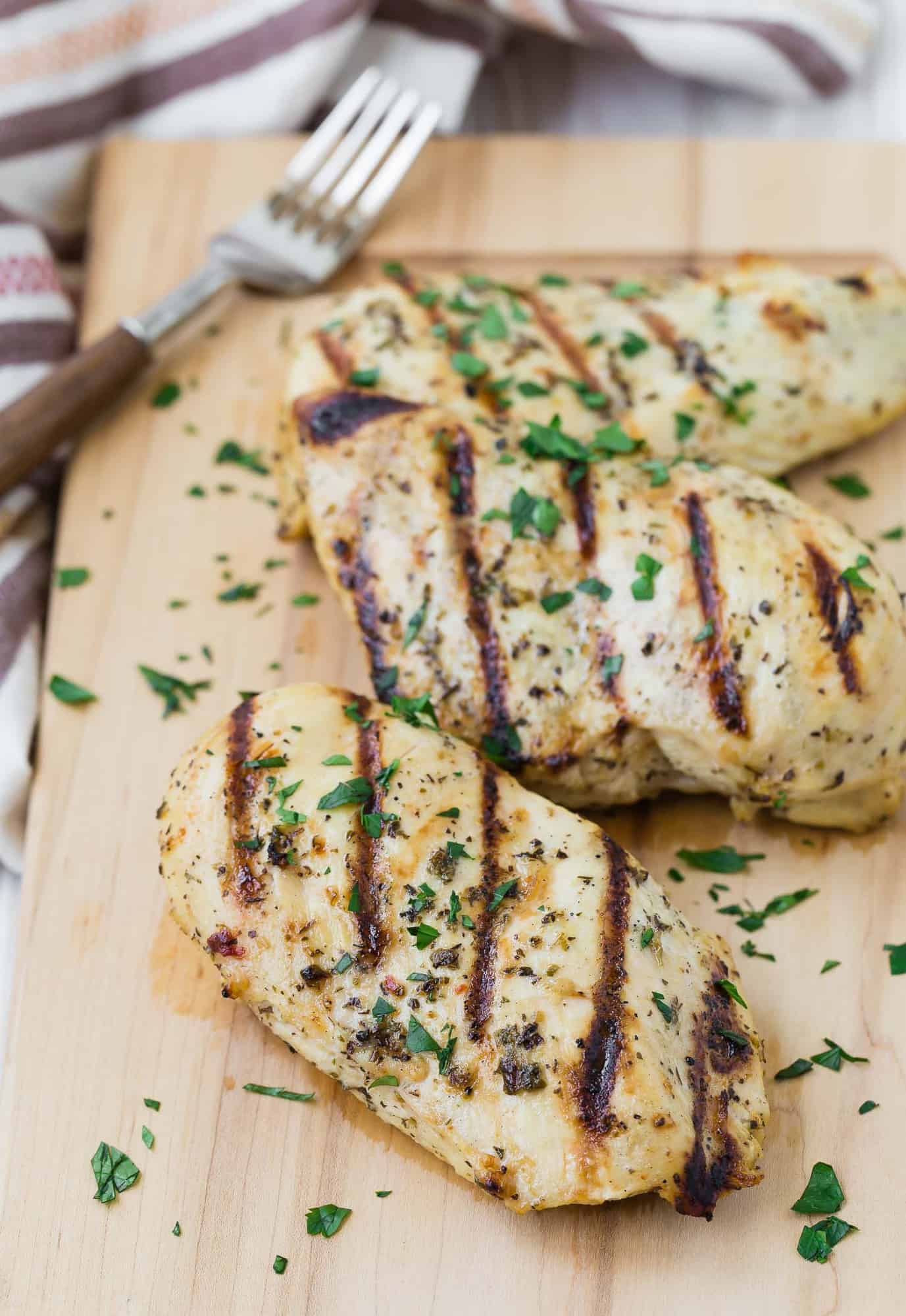 Three grilled boneless skinless chicken breasts on a wooden cutting board with a fork in the background.