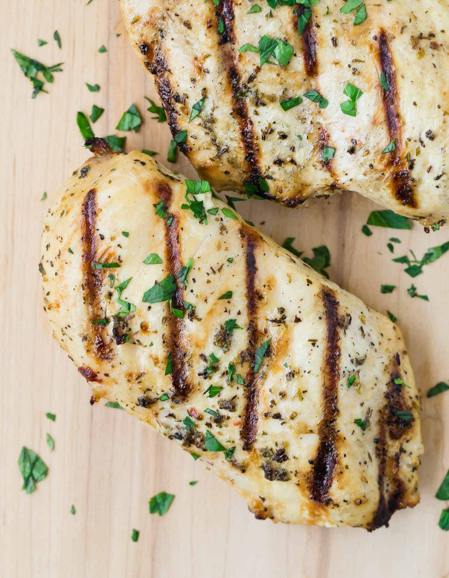 Overhead view of grilled chicken breasts on a wooden cutting board, sprinkled with fresh parsley.