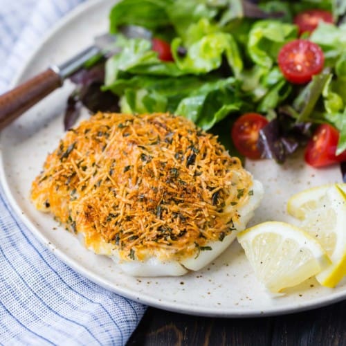 Parmesan baked cod on a plate with lemon wedges, a green salad, and a fork.