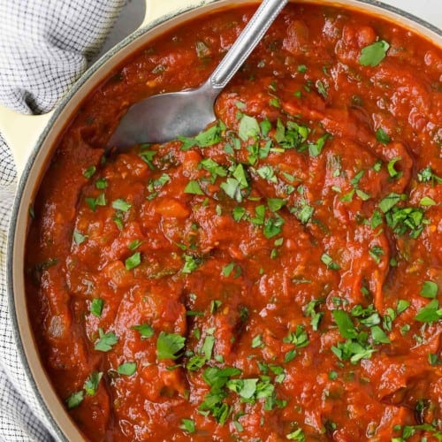 Image of a large pot of homemade spaghetti sauce with a spoon in it. It is garnished with lots of chopped fresh parsley.