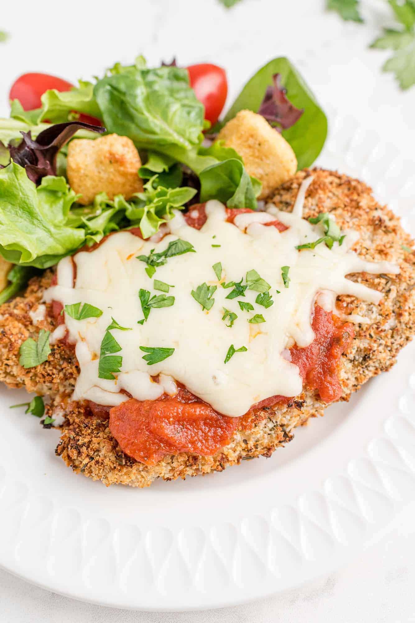 Baked chicken Parmesan on a white plate with a tossed green salad.