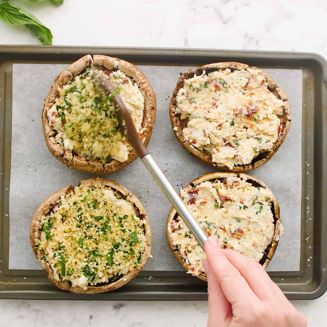 Breadcrumbs being spooned onto stuffed portobello mushrooms.