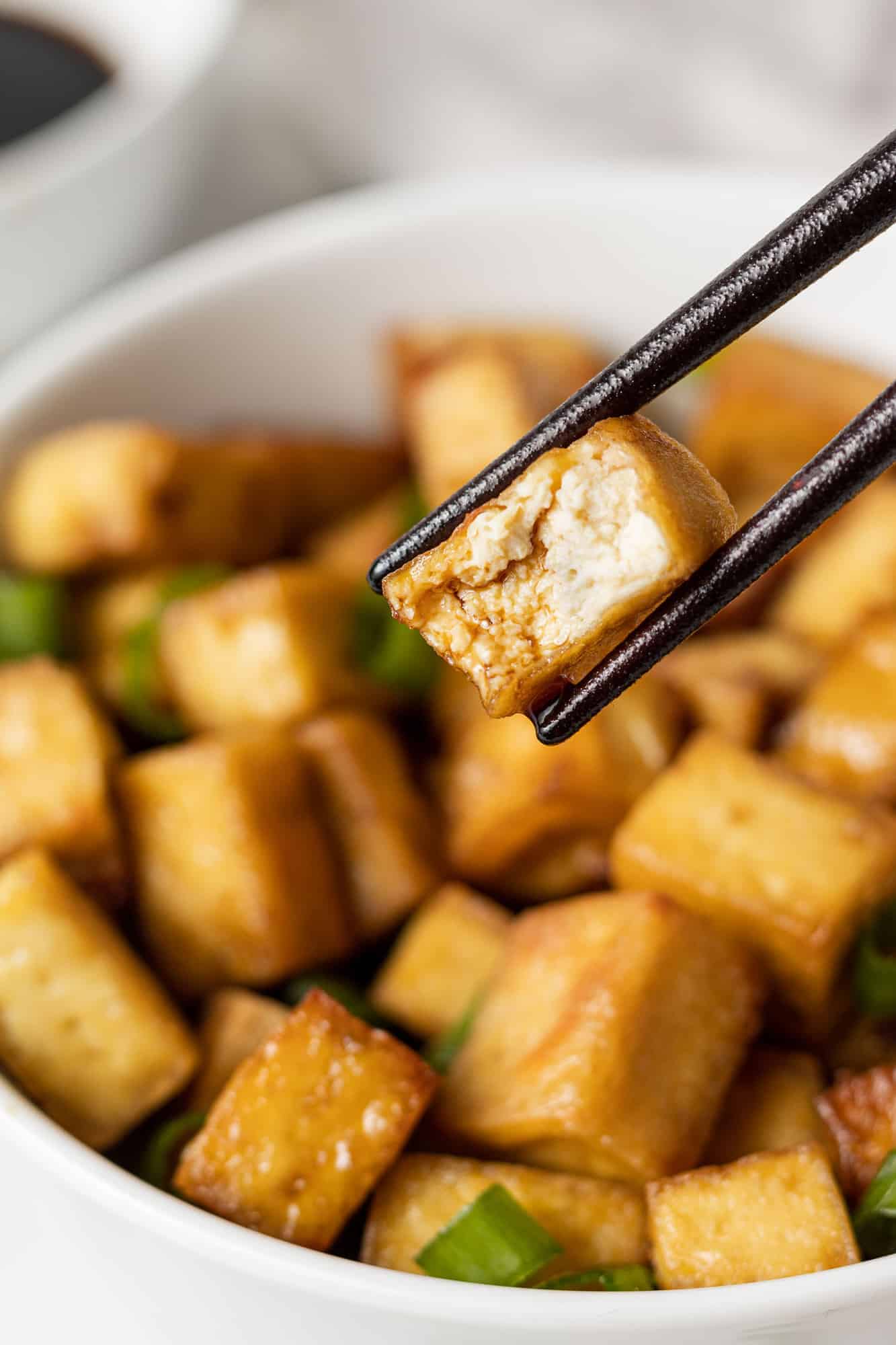 Tofu between chopsticks, more in a bowl in background.