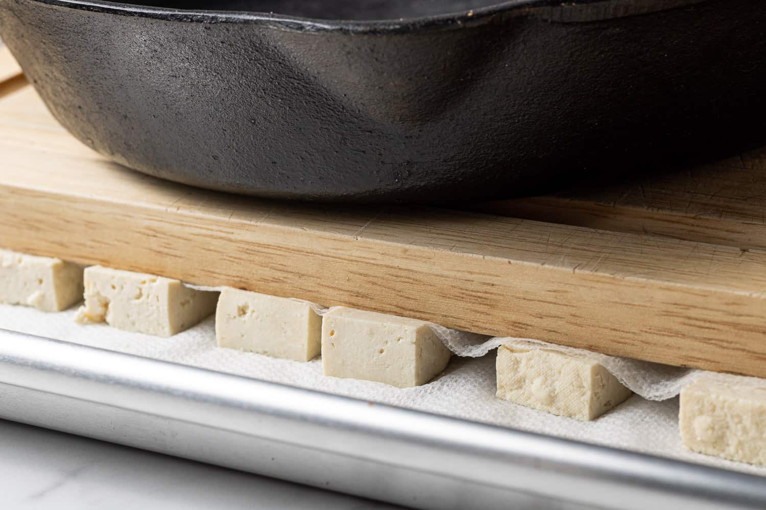 Tofu cubes on a sheet pan with paper towels, topped with cutting board and frying pan, showing how to press tofu.