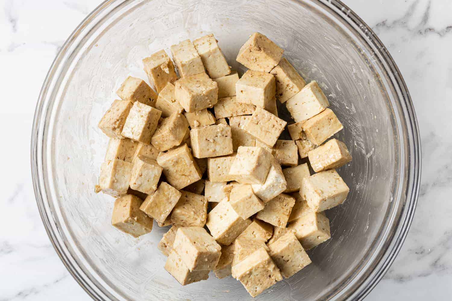 Tofu tossed in a clear glass bowl with soy sauce, oil, and cornstarch.