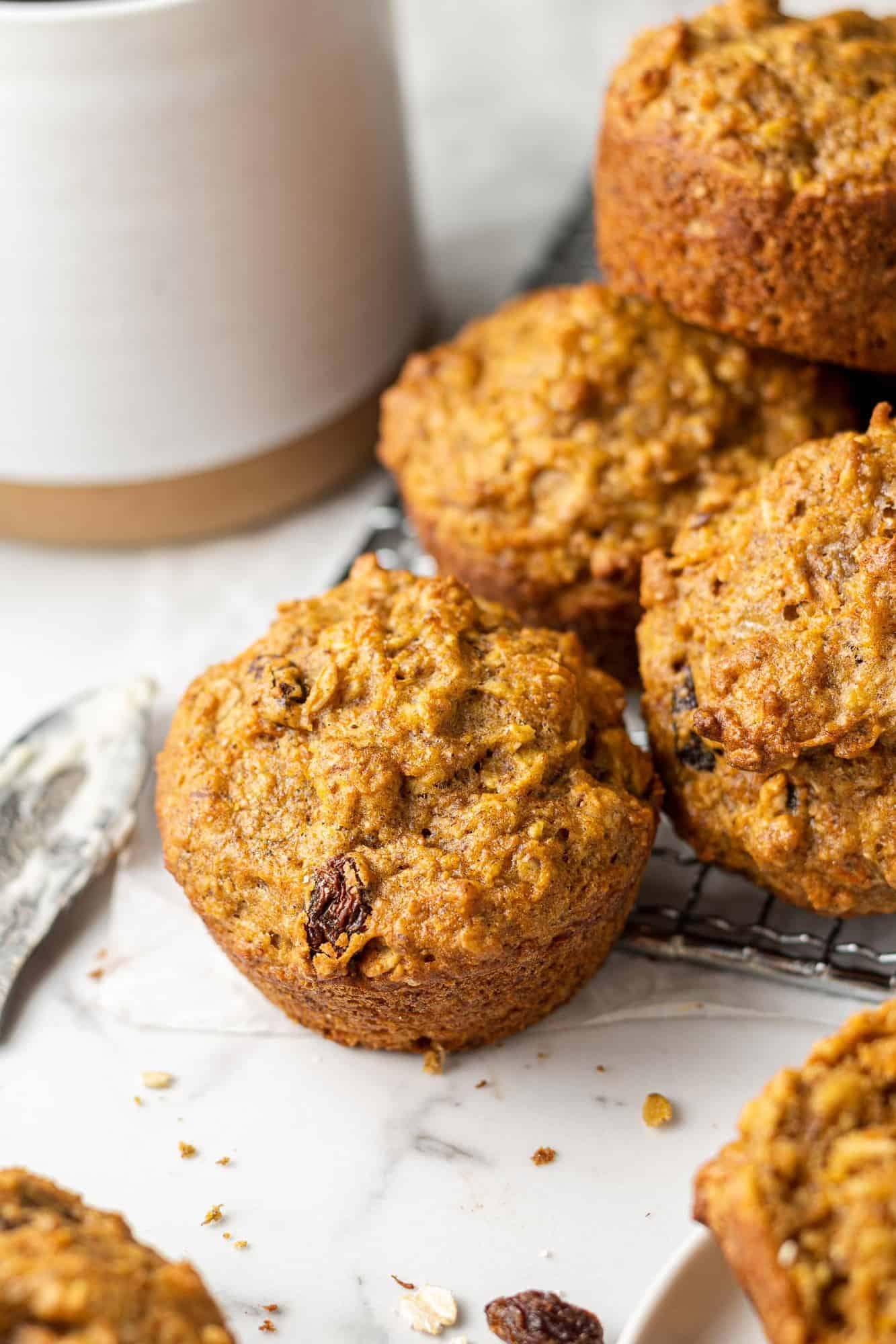 Pile of carrot raisin muffins on a white surface with a mug in the background.