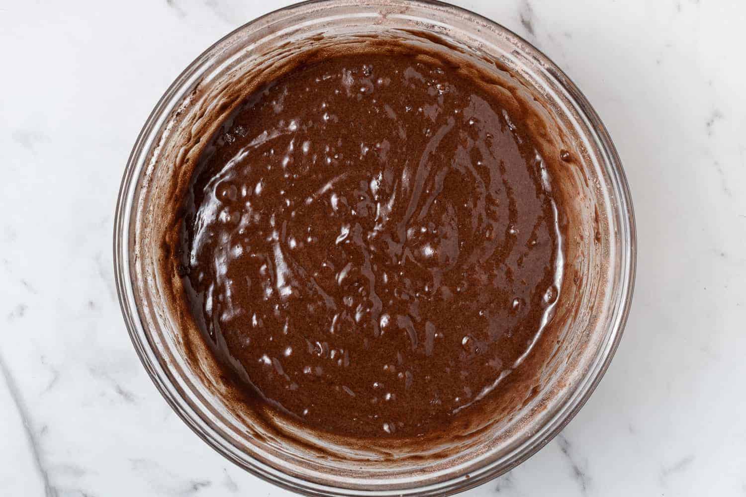 Chocolate cake batter in a glass mixing bowl on a marble surface.