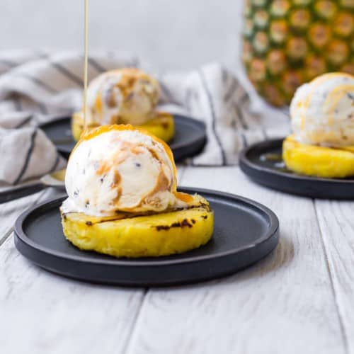 Grilled pineapple on a black plate, topped with ice cream, being drizzled with caramel.