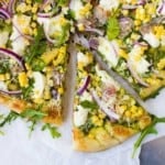 Overhead view of pizza topped with arugula, red onions, ham, and corn. One slice partially removed.