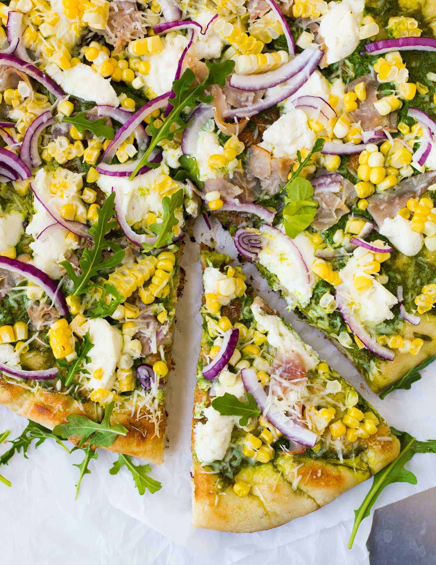 Overhead view of pizza topped with arugula, red onions, ham, and corn. One slice partially removed.