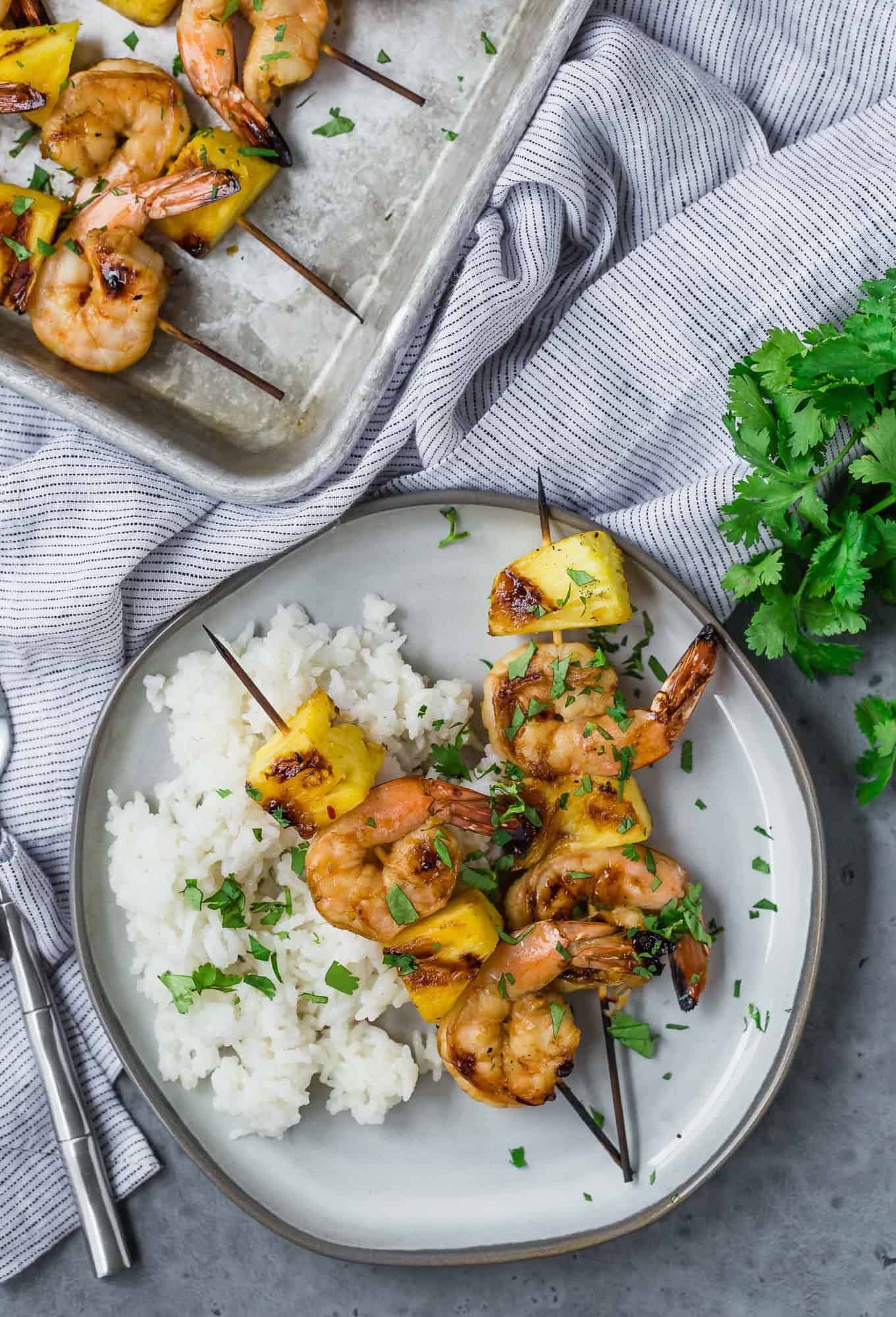 Overhead view of shrimp and pineapple, grilled on kabobs. Two kabobs are on a plate, and more are on a sheet pan nearby. Fresh cilantro and white rice is also pictured. 