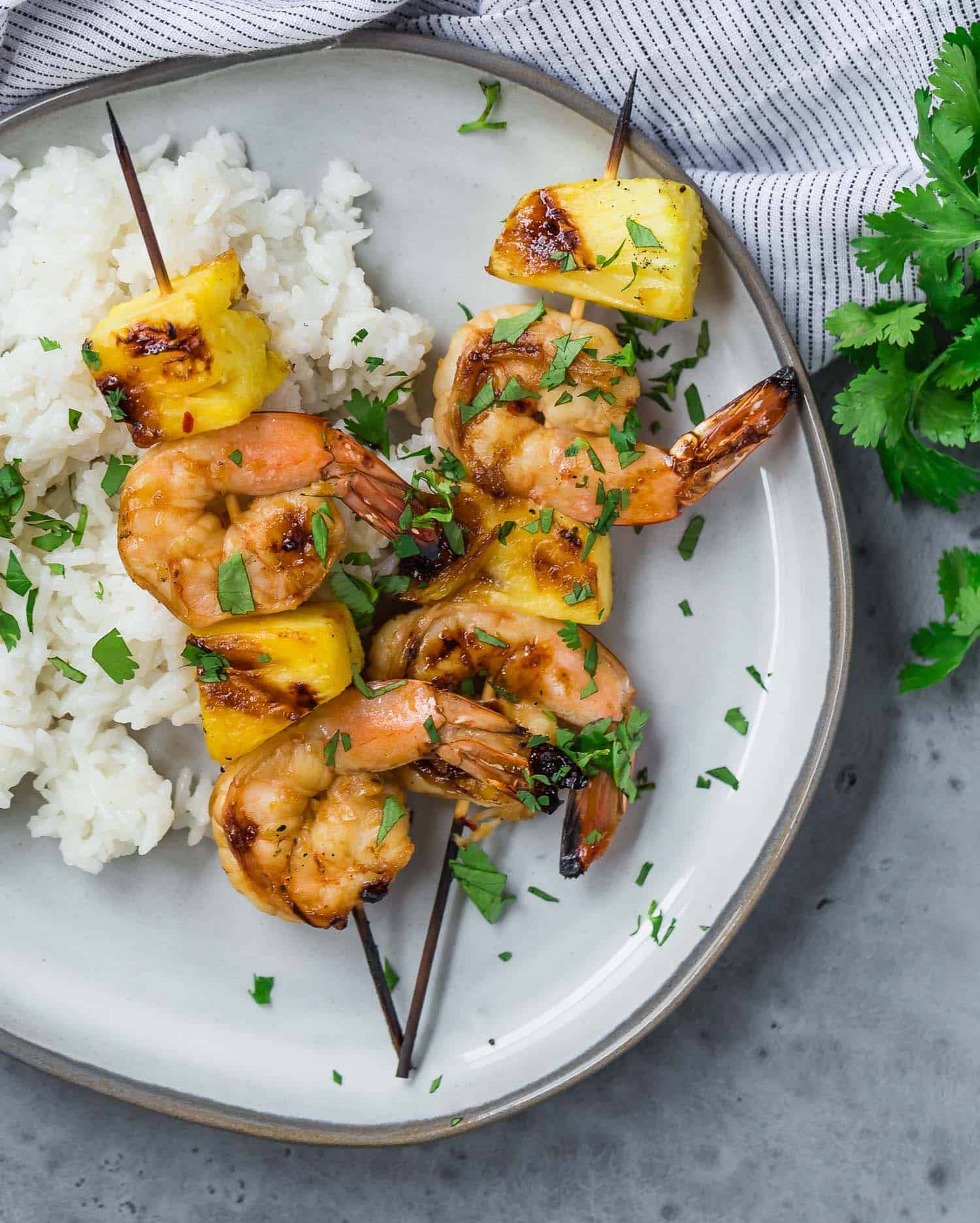 Overhead view of two grilled kabobs on a plate, with shrimp and fresh pineapple. They are served with white rice and sprinkled with fresh cilantro.