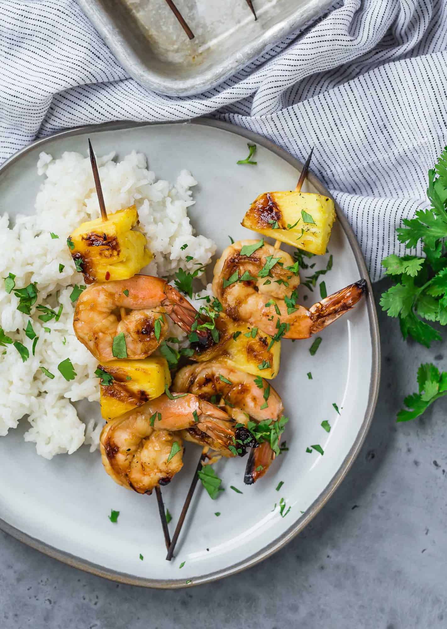 Overhead view of two grilled kabobs on a plate, with shrimp and fresh pineapple. They are served with white rice and sprinkled with fresh cilantro.