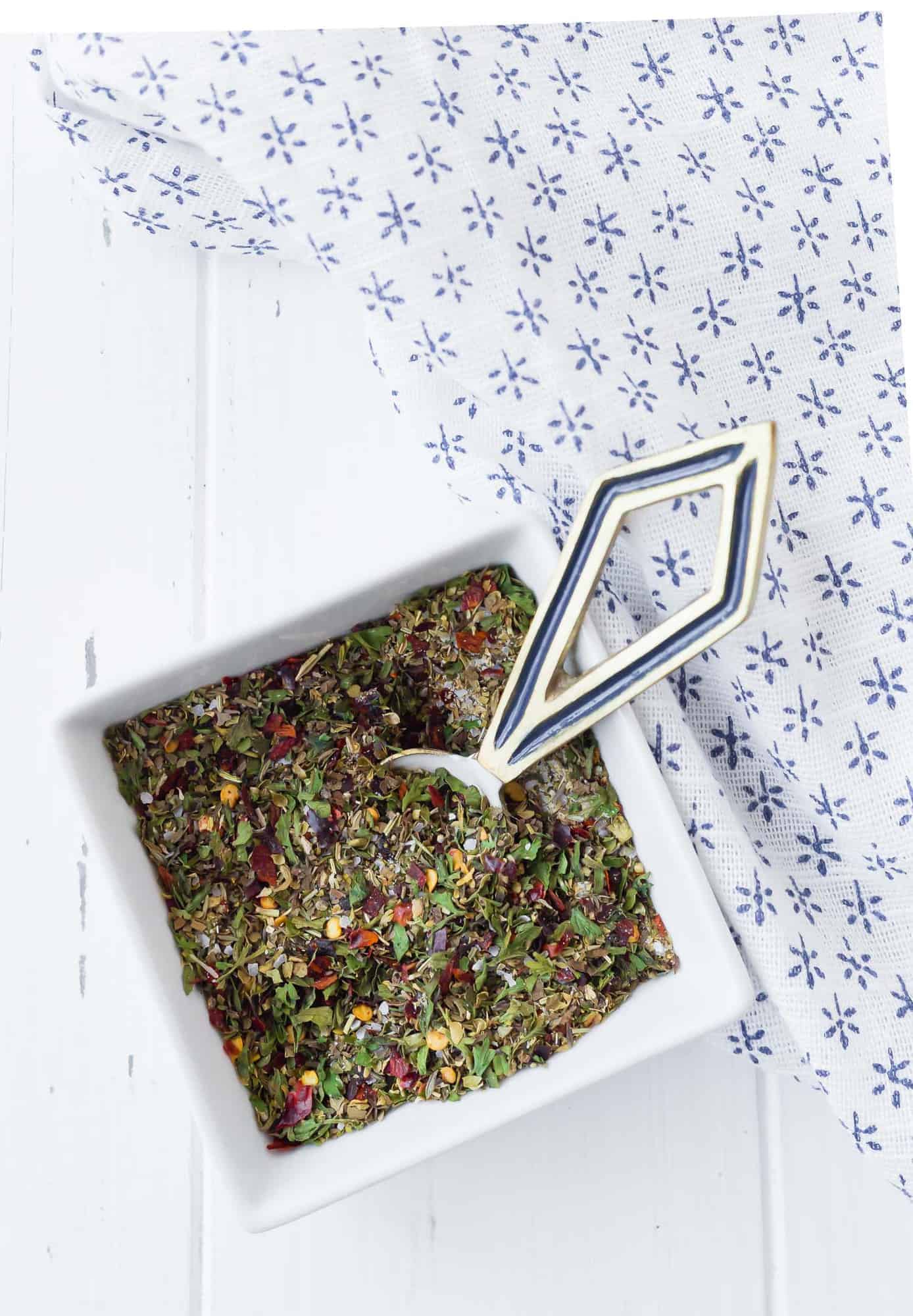 Dried herbs and spices in a small square white bowl with a decorative measuring spoon.