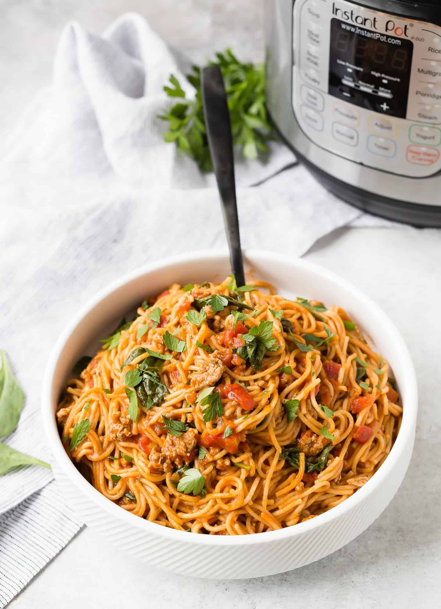 A large white bowl filled with spaghetti in tomato sauce, a pressure cooker and fresh parsley in the background of the image.