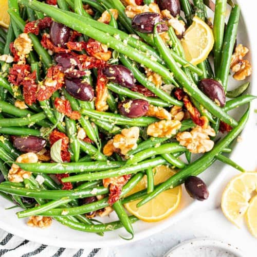 Green beans in a large white bowl with sun dried tomatoes, kalamata olives, and walnuts.