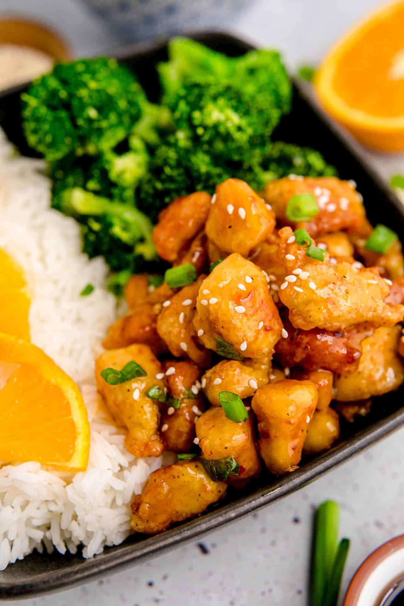 Homemade orange chicken on a black plate with rice and broccoli.