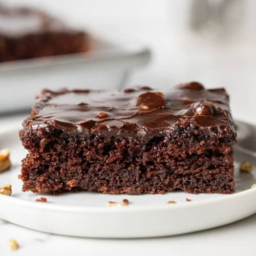 Square piece of chocolate cake with thin frosting, on a white plate.