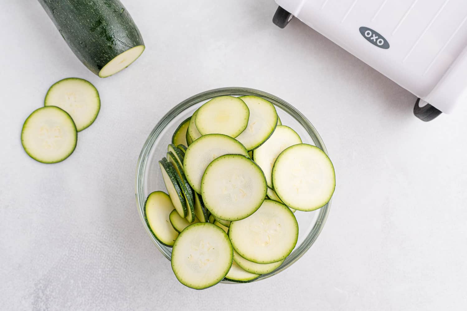Sliced zucchini in a bowl, a whole zucchini partially visible, a mandoline in the upper right hand corner.
