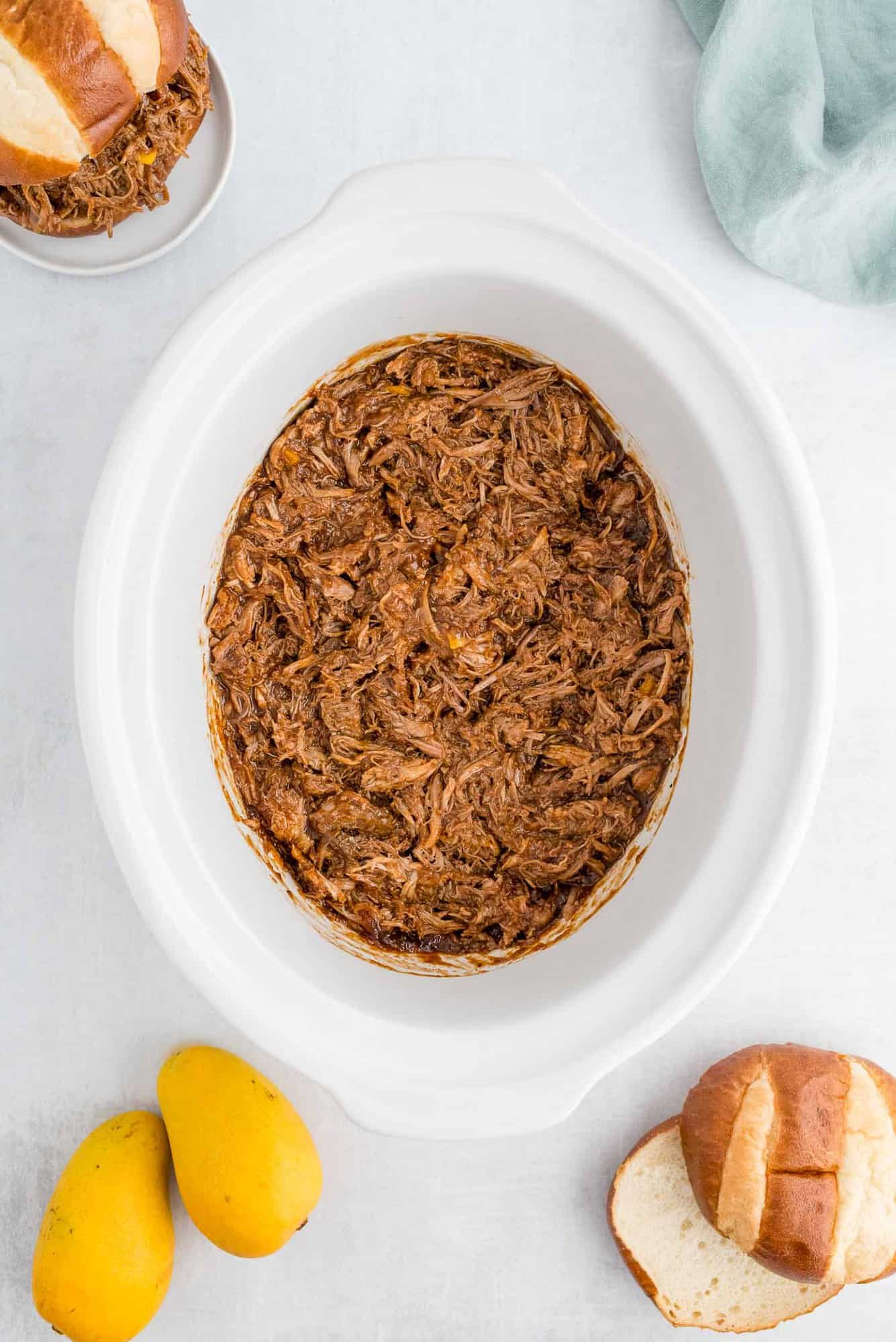 Pork in a slow cooker surrounded by mangos and pretzel rolls.