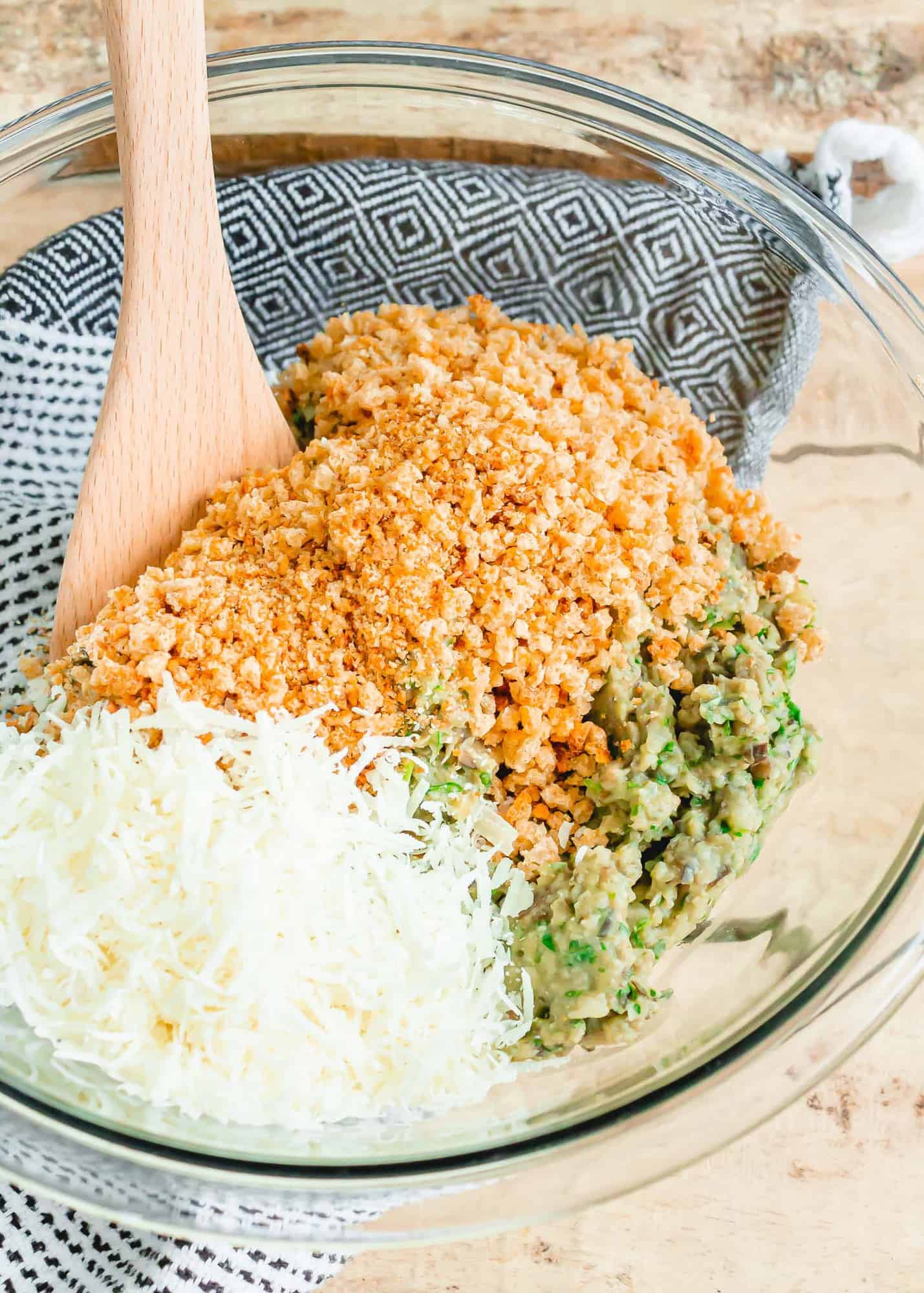 Breadcrumbs and cheese in a clear glass bowl with a wooden spoon.