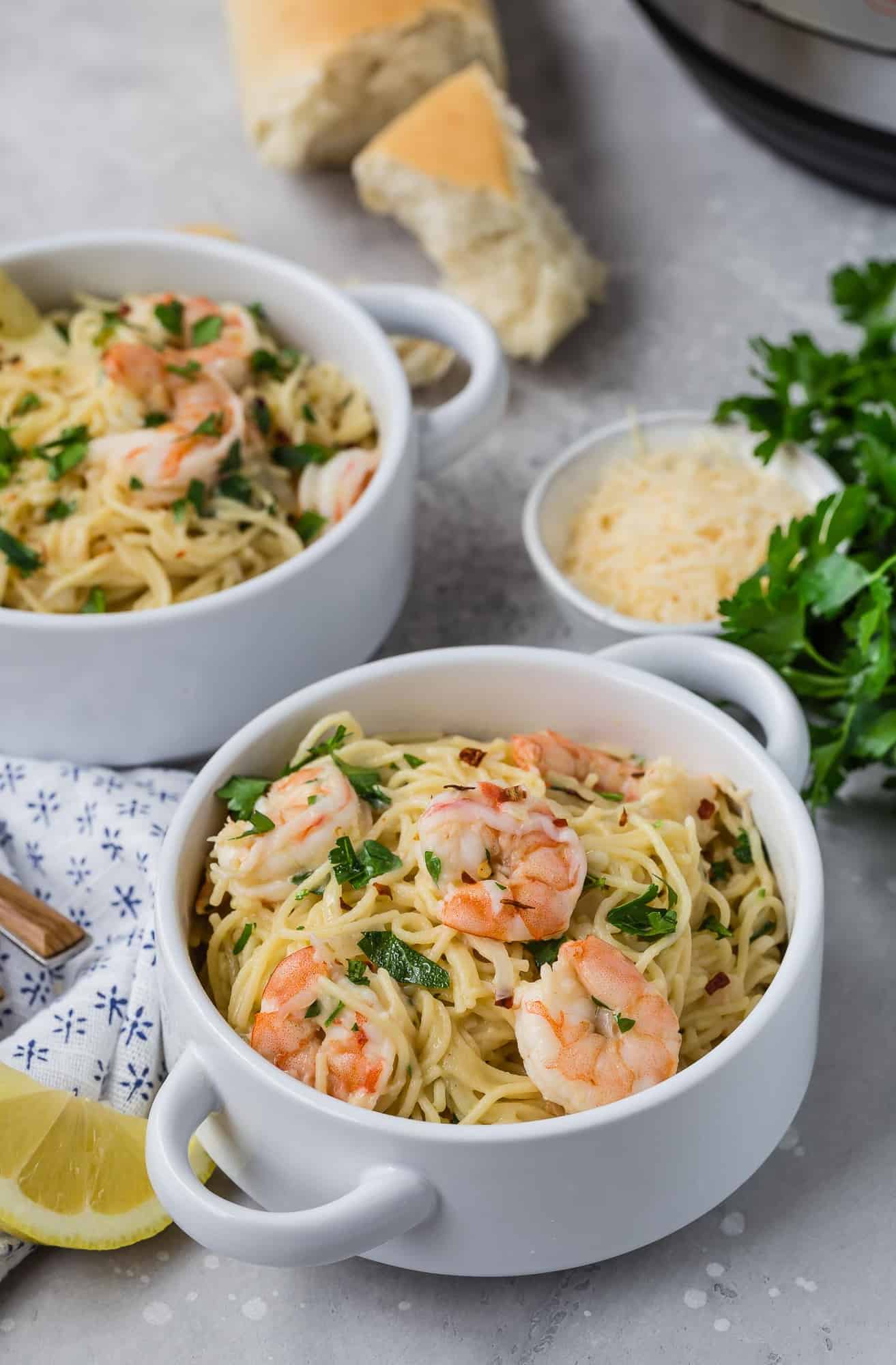 Two bowls of shrimp scampi pasta, bread, cheese, and parsley in background.