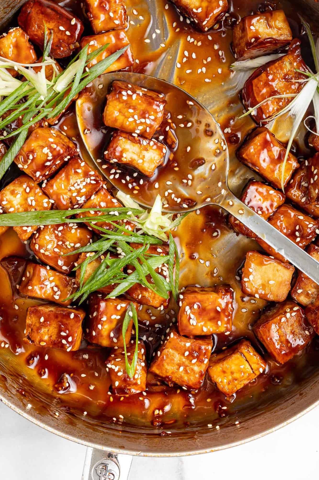 Overhead view of sticky sesame tofu in a frying pan with green onions.