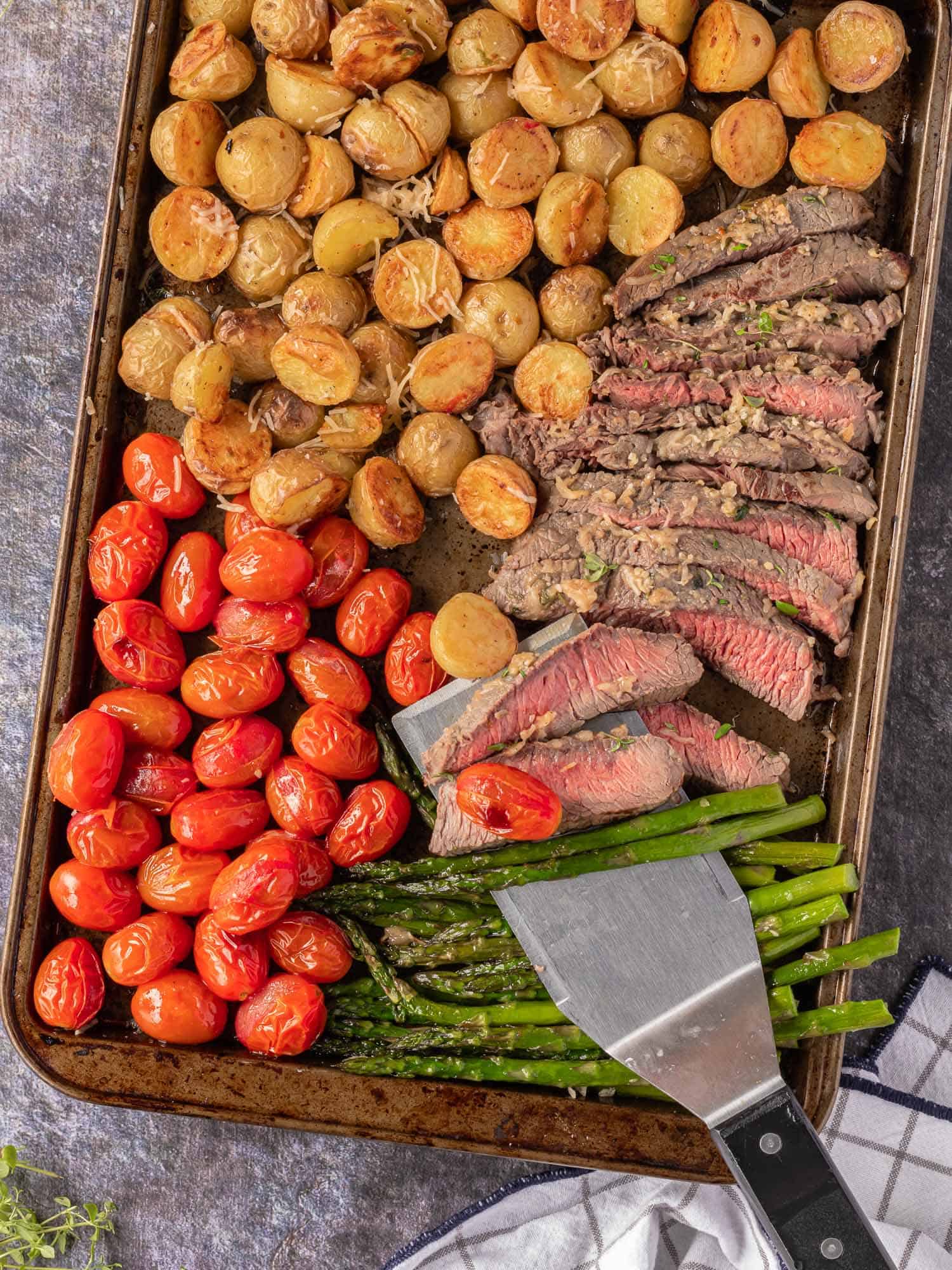 Steak on a spatula, surrounded by other food: tomatoes, asparagus, and potatoes.