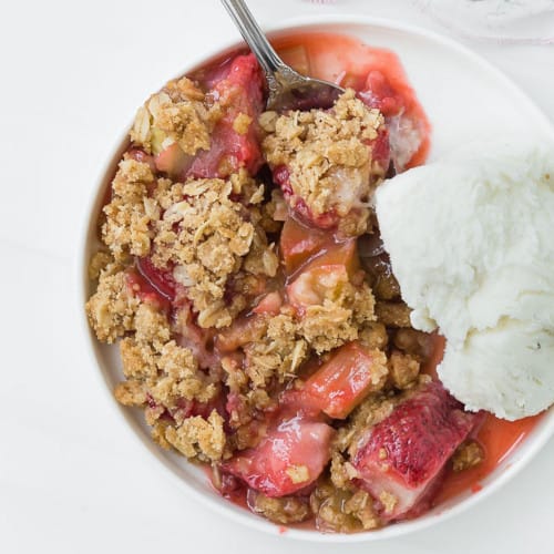 Close up view of strawberry rhubarb crisp and vanilla ice cream on a round plate, with a spoon.