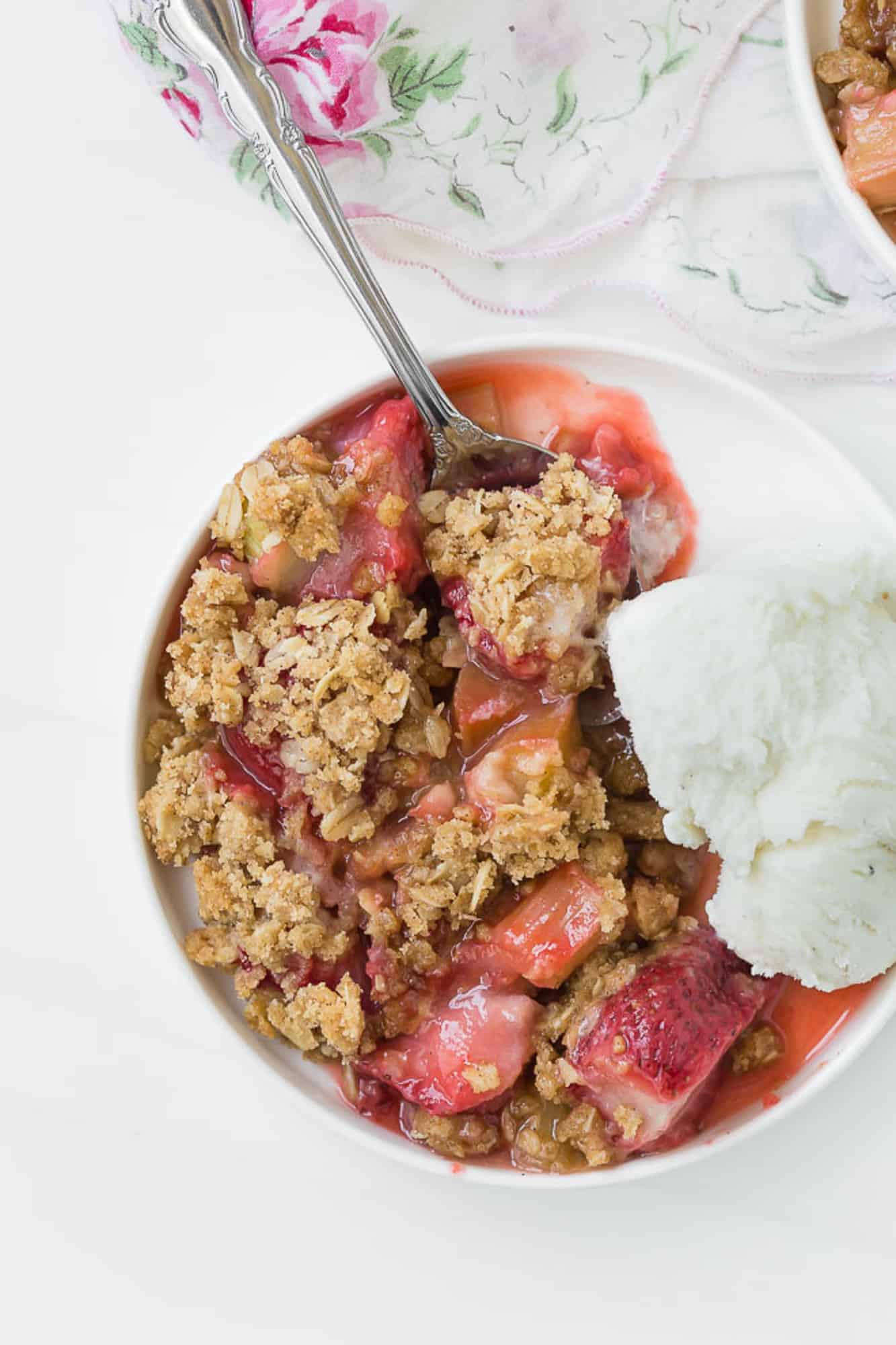 Close up view of strawberry rhubarb crisp and vanilla ice cream on a round plate, with a spoon.