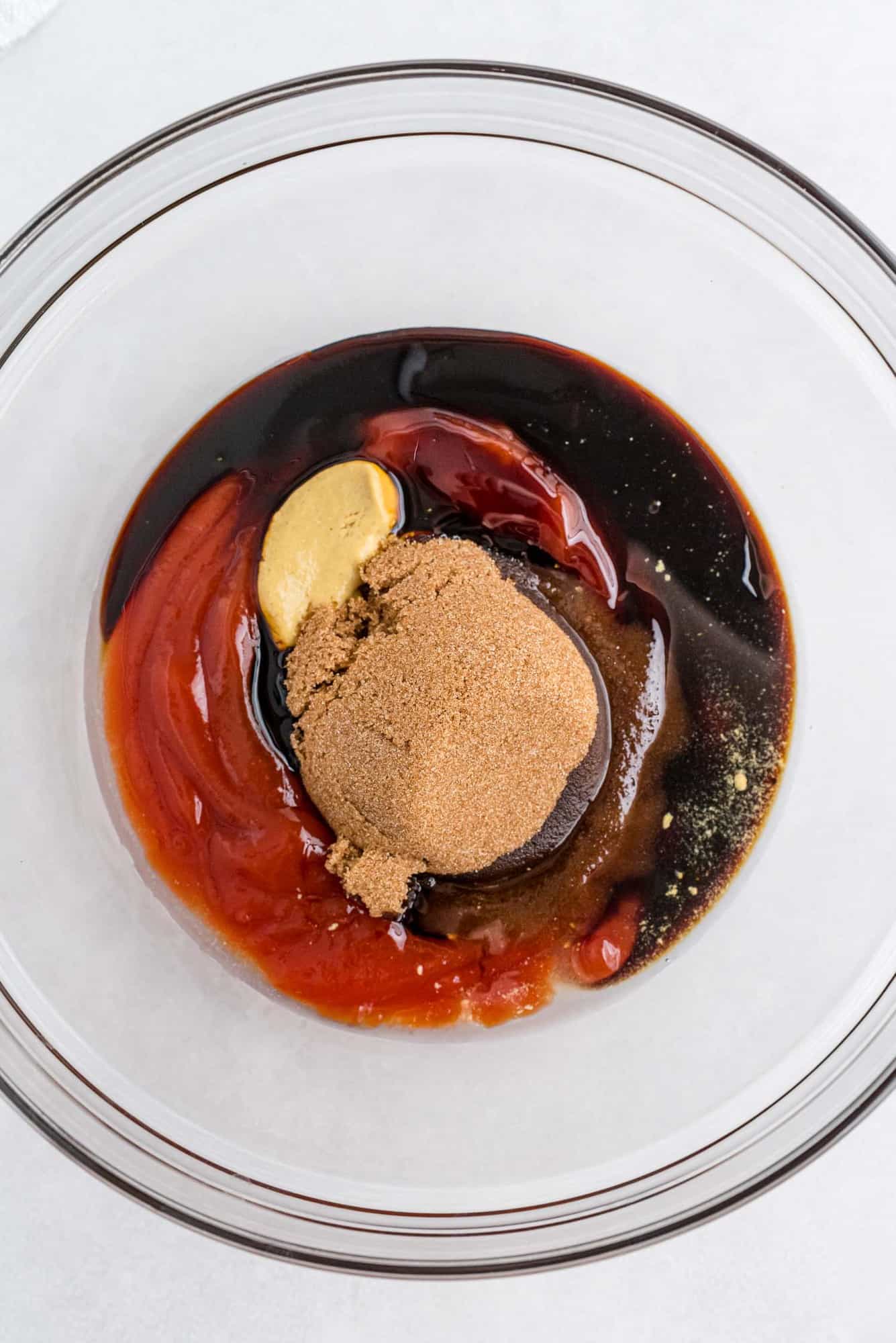 A glass bowl containing brown sugar, ketchup, mustard, and molasses before being mixed together.