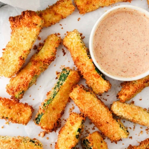 Air fryer zucchini fries on parchment with a light red dipping sauce.