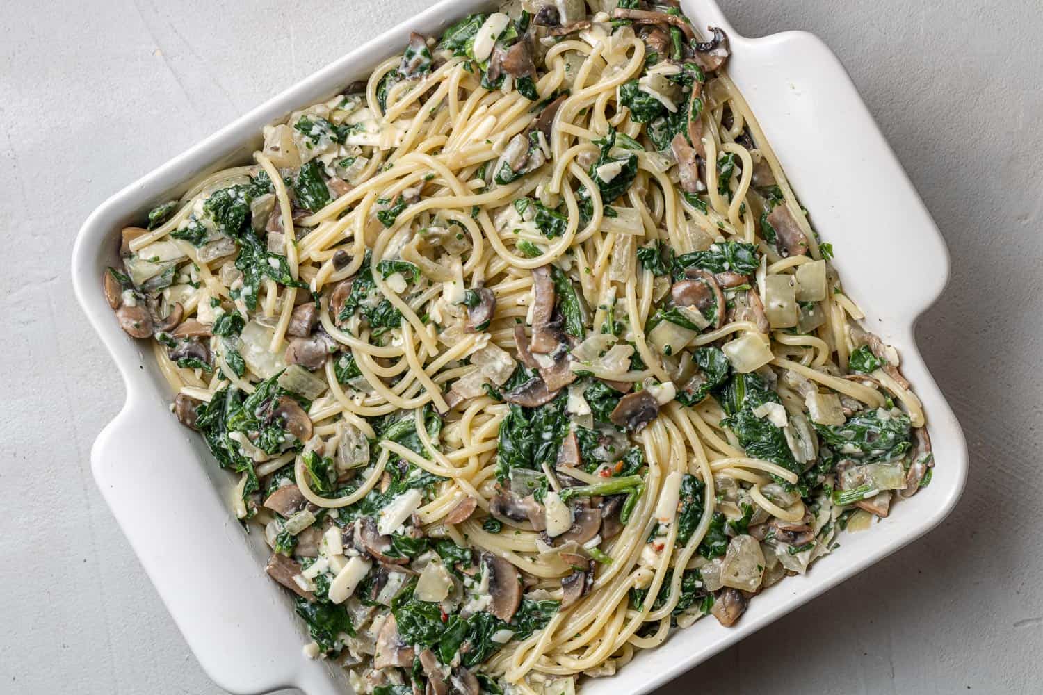 Unbaked spaghetti with spinach and mushrooms in a square white baking dish.