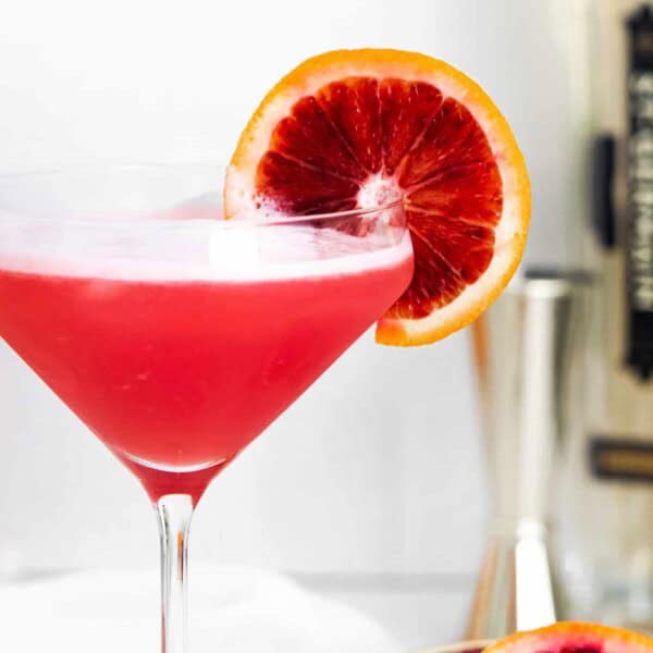 Close up of martini glass with bright red cocktail garnished with blood orange.