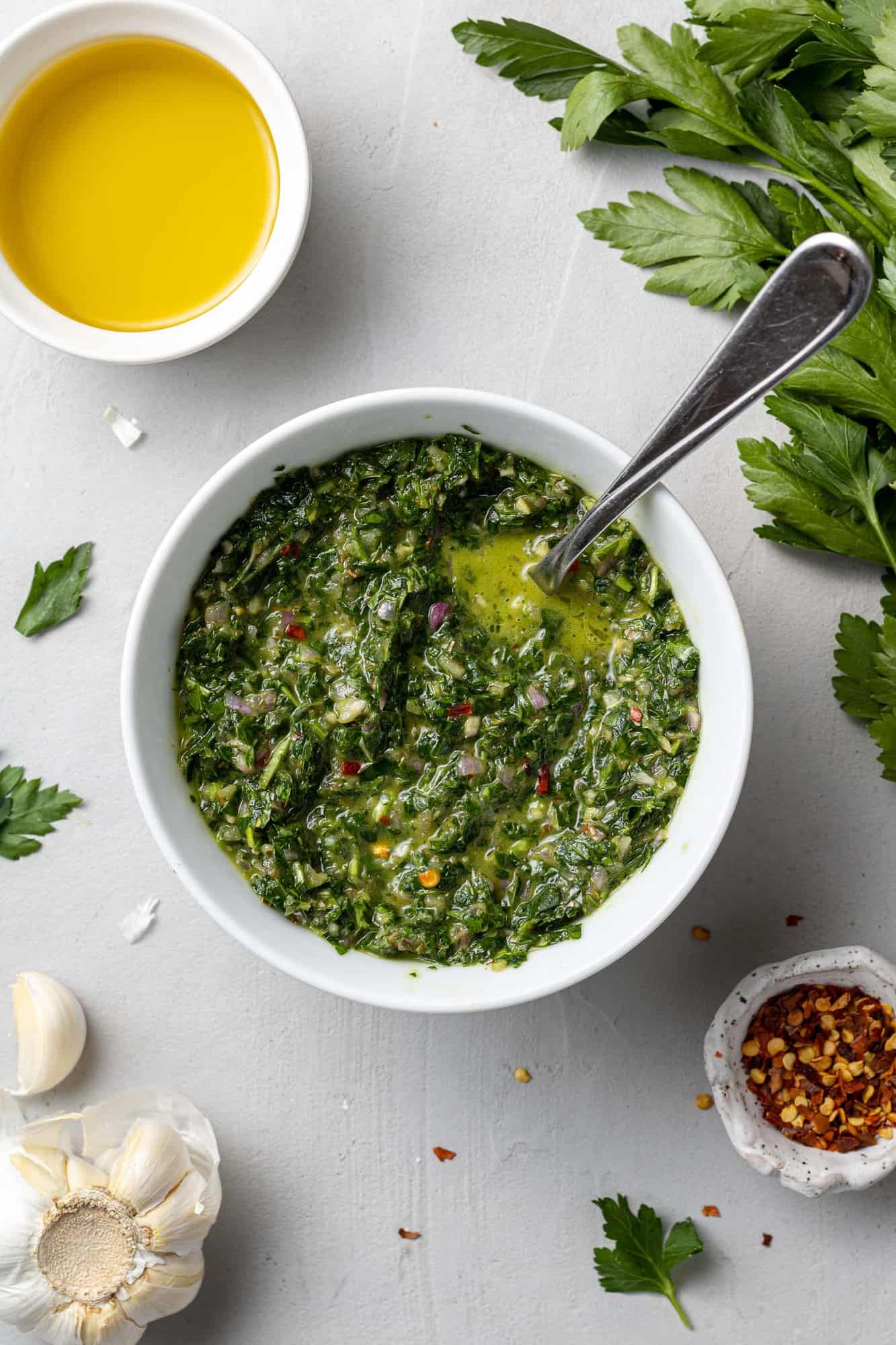 Overhead view of chimichurri sauce in a white bowl, surrounded by ingredients.