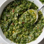 Green chimichurri sauce in a white bowl, a spoon lifting some up to show texture.