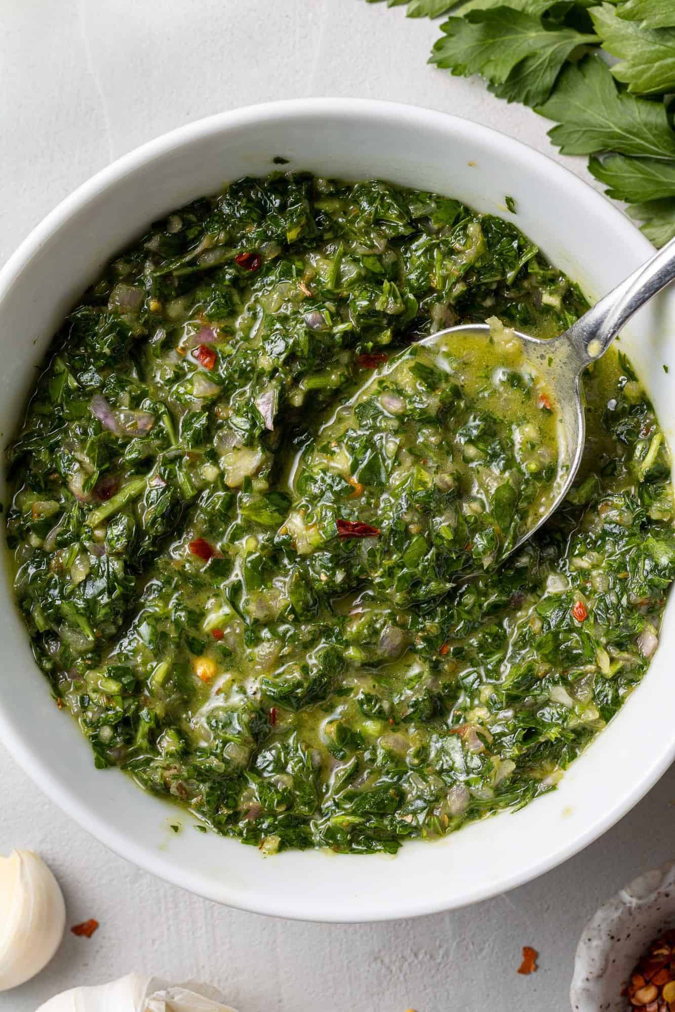 Close up of chimichurri sauce in a white bowl with a silver spoon.