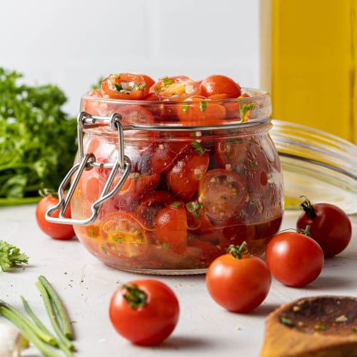 Tomatoes with olive oil and herbs in a small glass jar. Fresh tomatoes in the foreground.