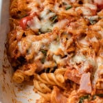 Pasta in a baking dish with pizza sauce, ham, cheese, tomatoes, and spinach. A scoop has been removed.