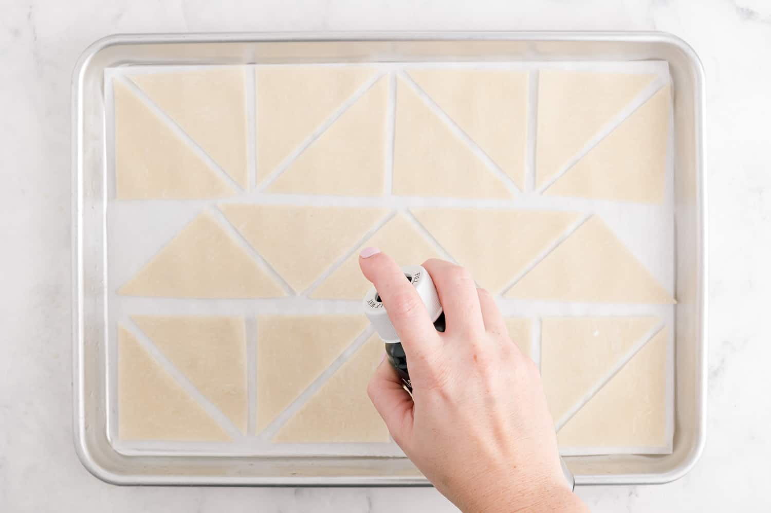 Wonton wrappers being sprayed with oil on a sheet pan.