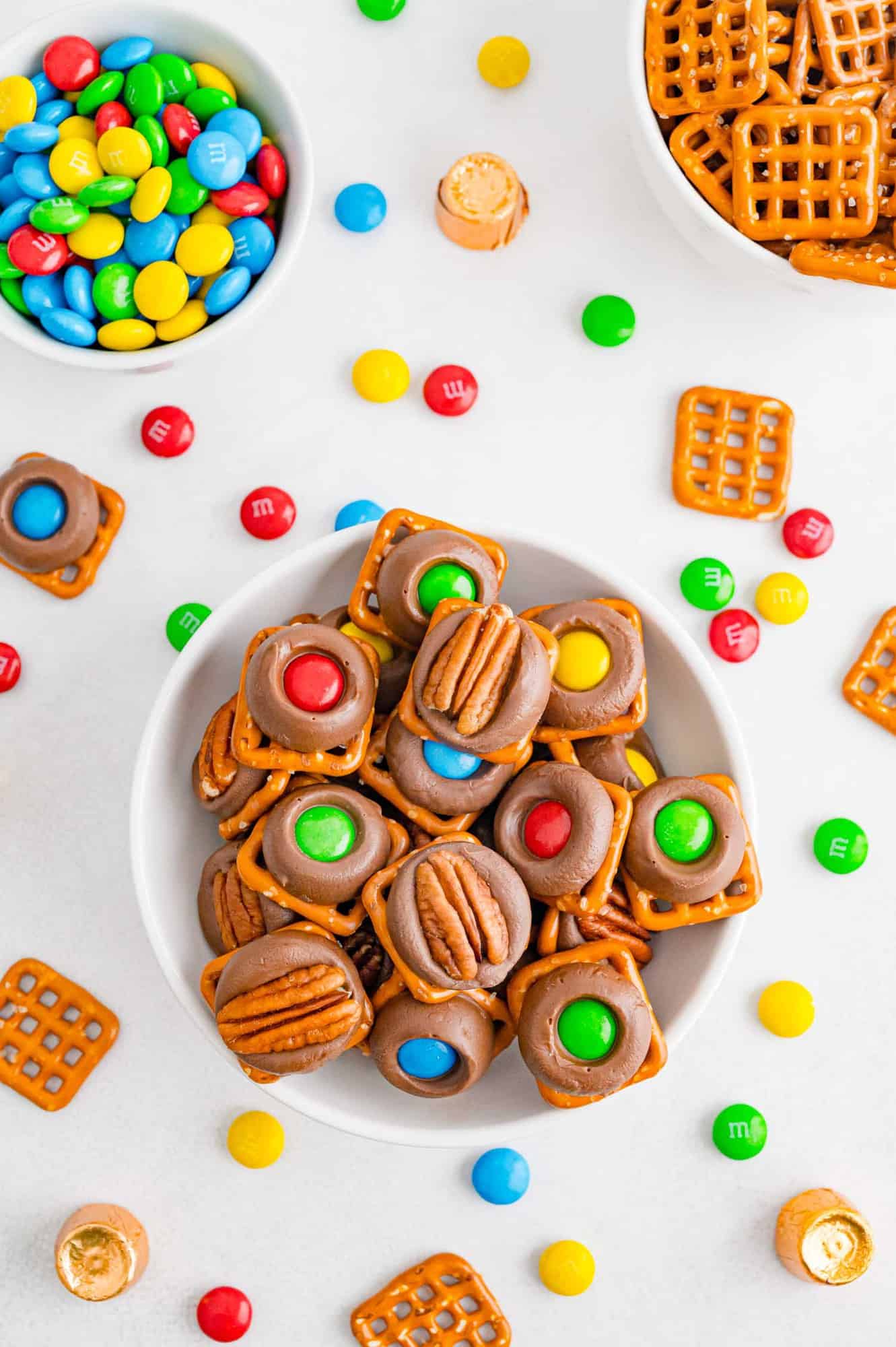 Bowl of rolo pretzels topped with pecan halves and colorful M&Ms.