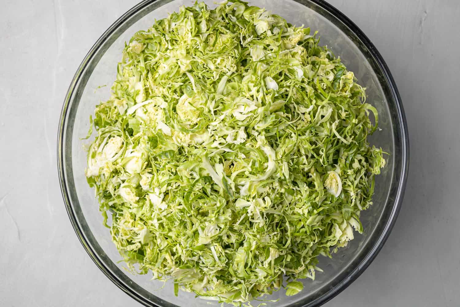 Shredded Brussels sprouts in a large glass bowl on grey surface.