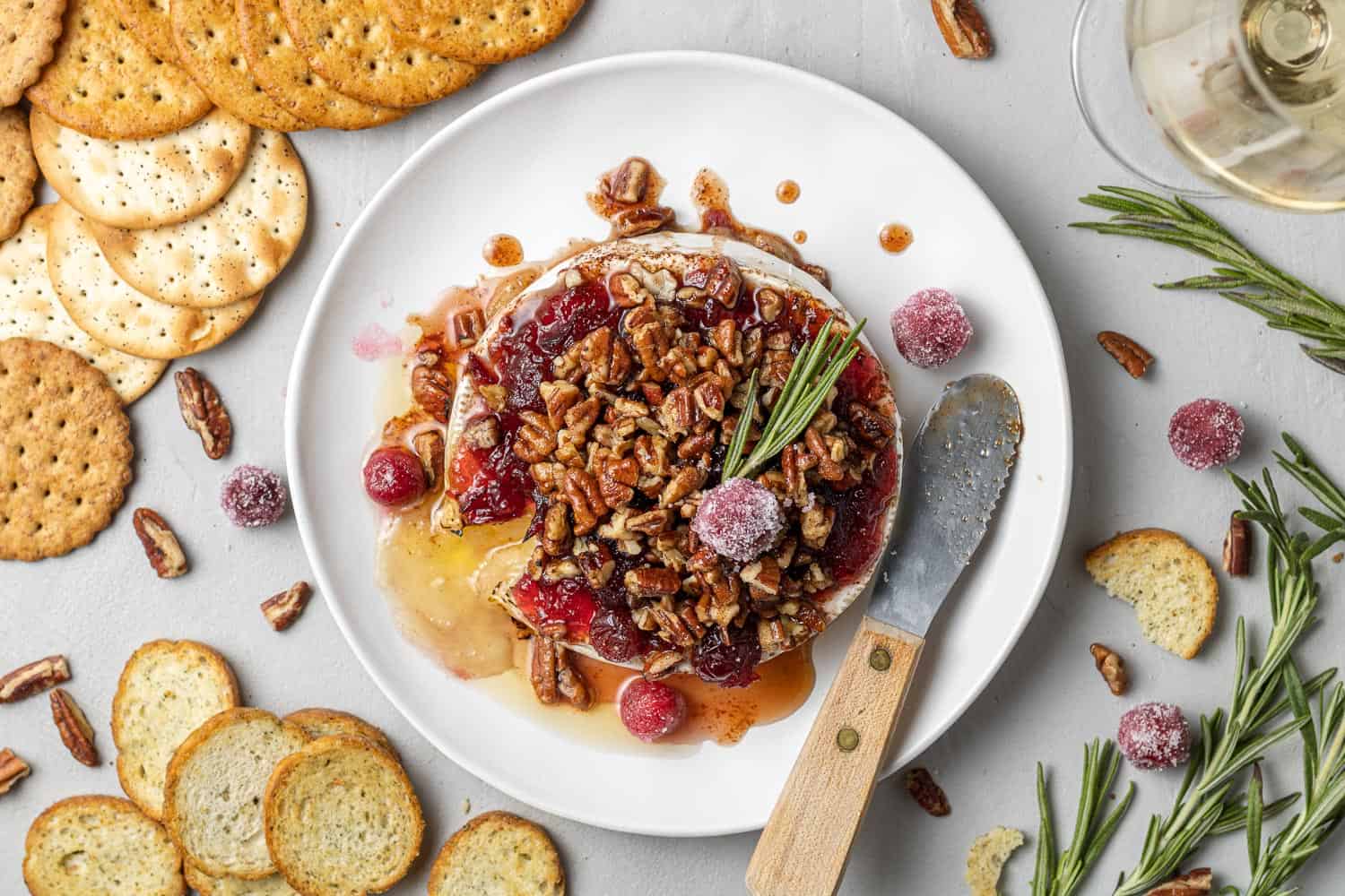 Baked brie on a plate surrounded by crackers, rosemary, and candied cranberries.