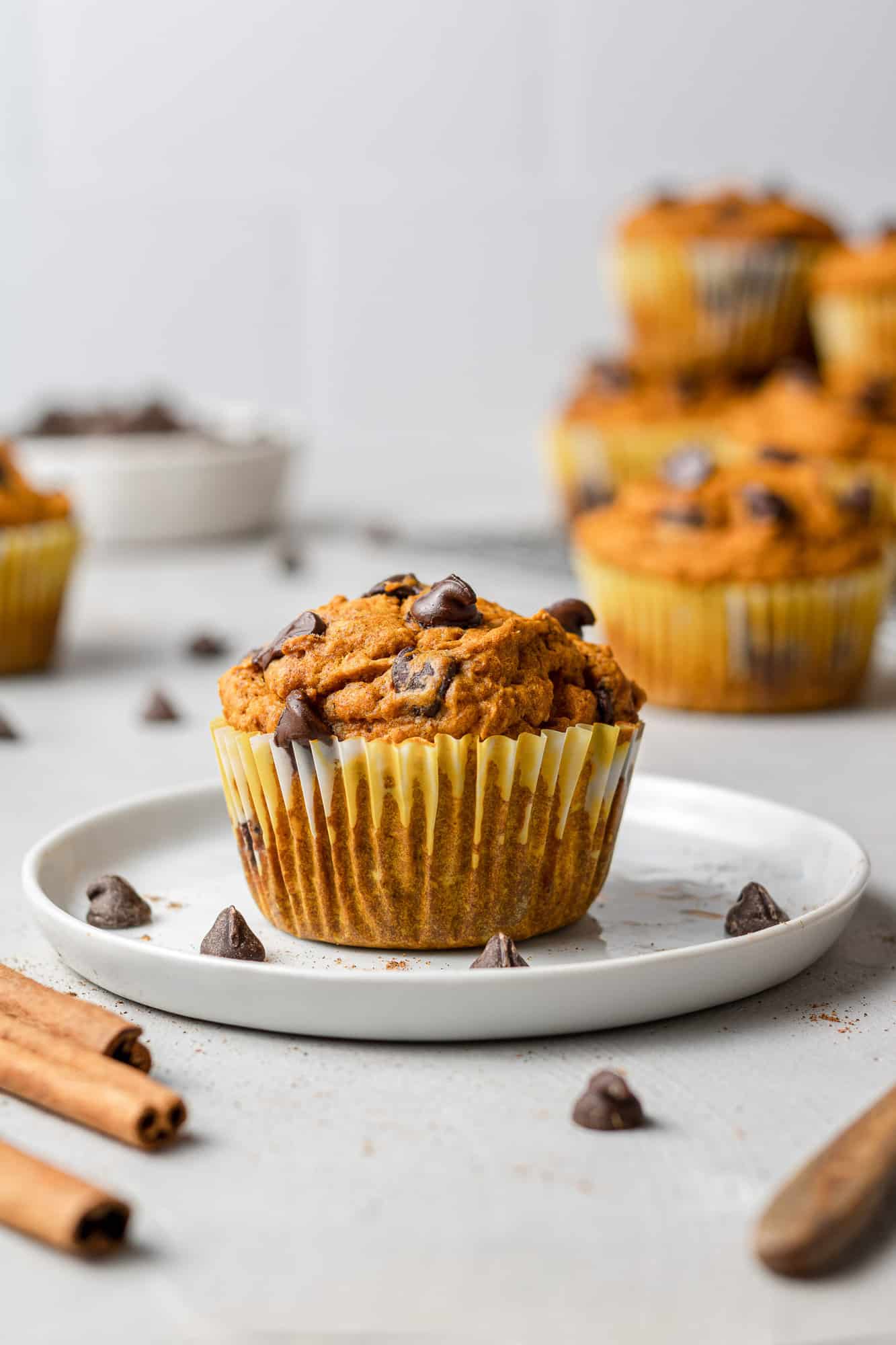 One pumpkin chocolate chip muffin on a small white plate, more muffins in the background.