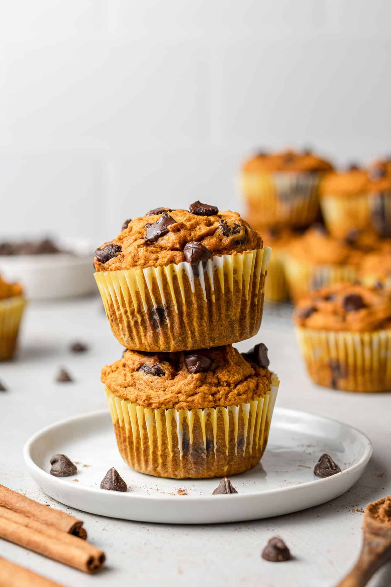 Two pumpkin chocolate chip muffins stacked on a small white plate, with more muffins in the background.