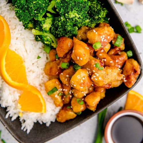 Orange chicken on a black plate with broccoli and rice.