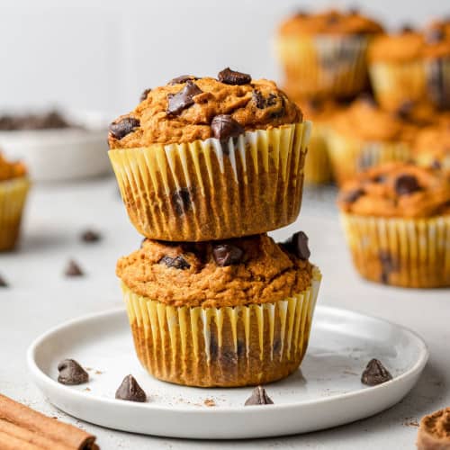 Two pumpkin chocolate chip muffins stacked on a small white plate, with more muffins in the background.