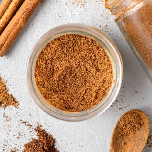 Overhead view of pumpkin pie spice in a small jar, cinnamon sticks nearby.